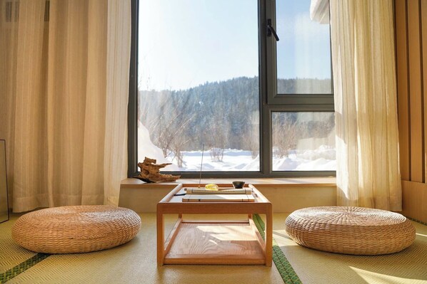A room with a large window, two woven poufs, a wooden table with a bowl and a spoon, and a piece of driftwood.