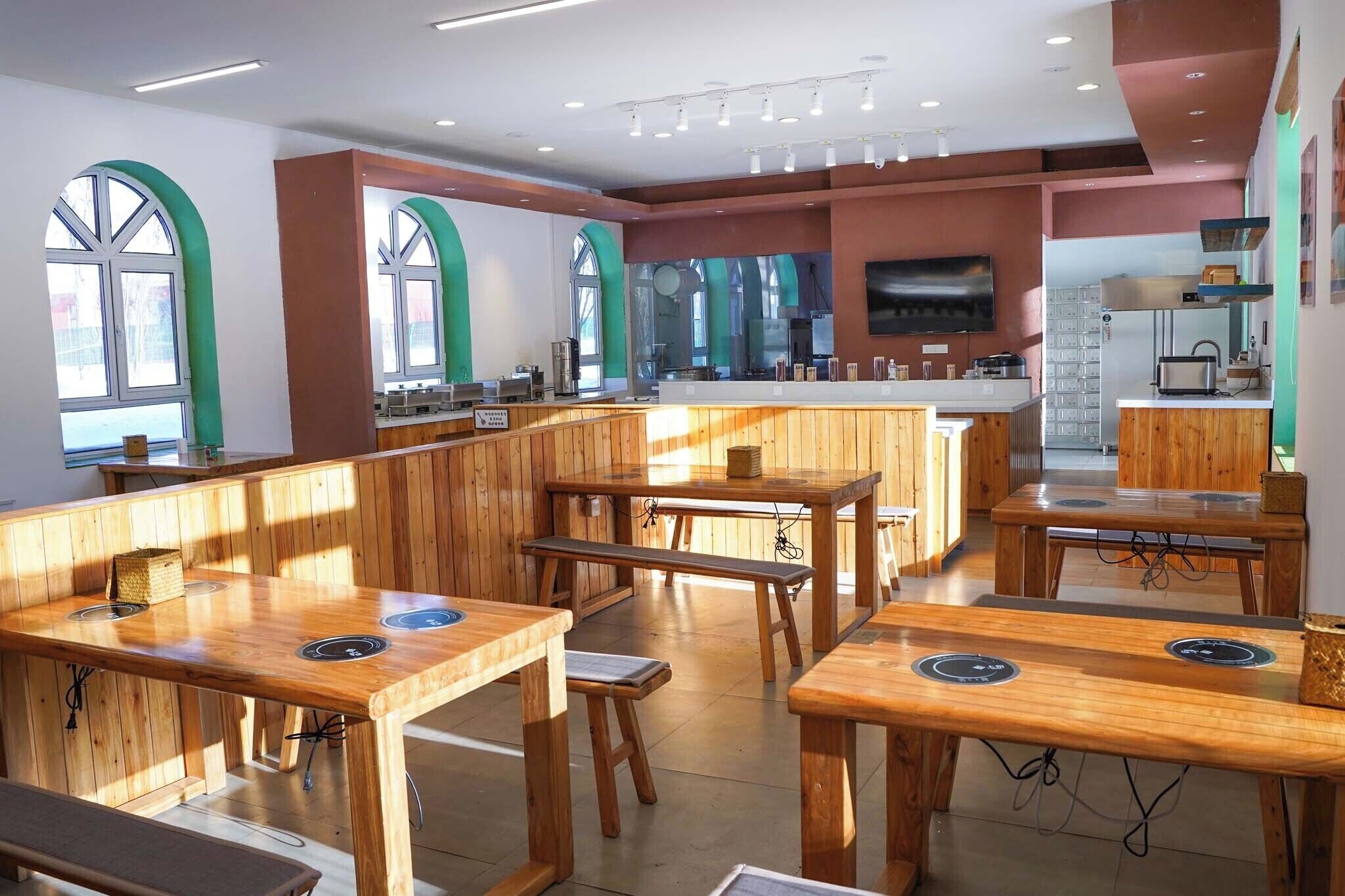 Un restaurante con mesas y bancos de madera, una zona de cocina y grandes ventanales.