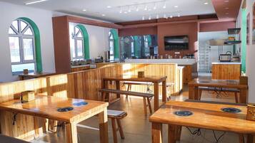 Un restaurante con mesas y bancos de madera, una zona de cocina y grandes ventanales.