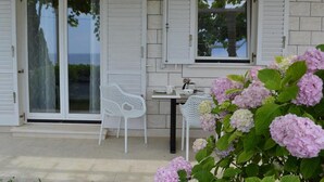 Ein Haus mit einer Terrasse, weißen Fensterläden, einem Tisch mit Stühlen und einem Garten mit rosa Blumen.