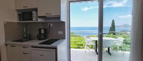 Une cuisine compacte équipée d’un four à micro-ondes, d’une machine à café et d’un lave-vaisselle Monocar. Adjacente à la cuisine, une porte-fenêtre coulissante donne accès à un espace extérieur aménagé avec une table et des chaises, offrant une vue panoramique sur la mer et la verdure.