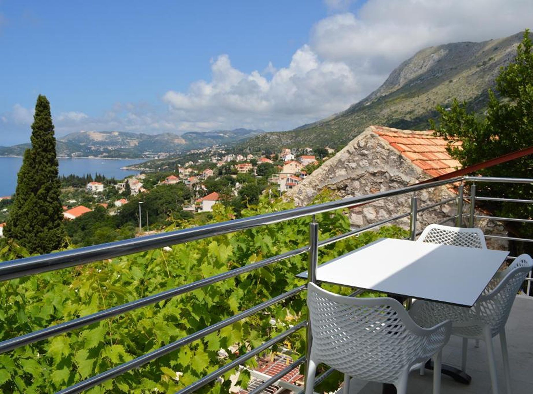 Een balkon met tafel en stoelen, uitkijkend op een kustplaatsje met een berg op de achtergrond.