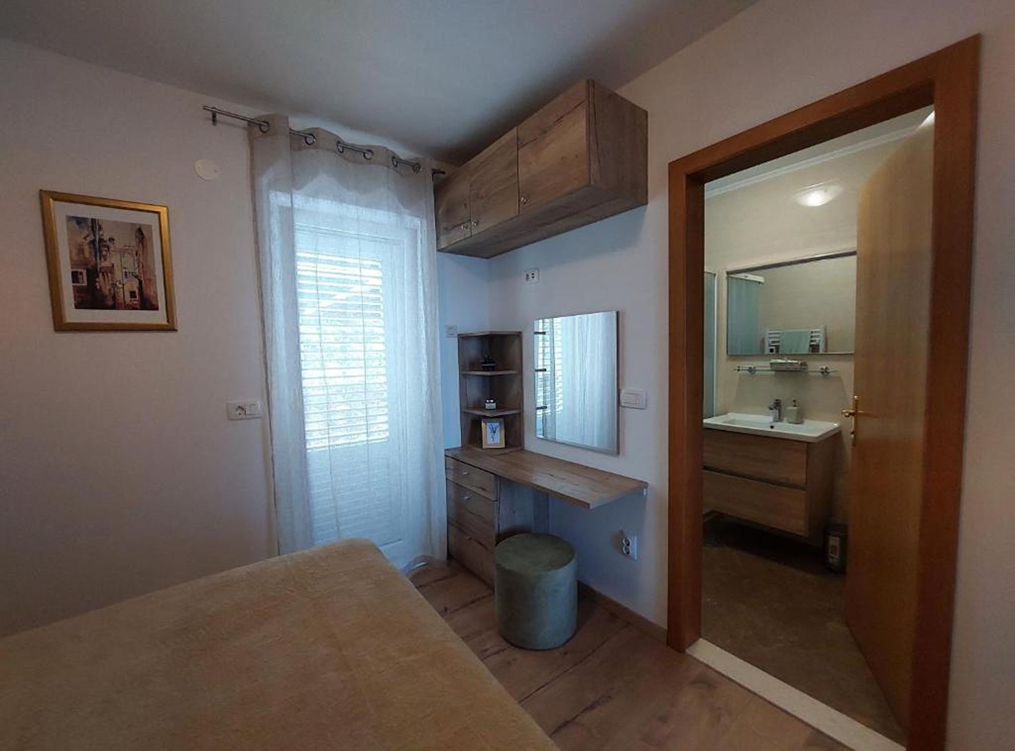 A bedroom with a bed, a desk, a chair, a small trash bin, a window with curtains, and a bathroom with a mirror and sink.