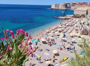 Una playa concurrida con sombrillas y gente, un mar azul cristalino y un edificio histórico al fondo.