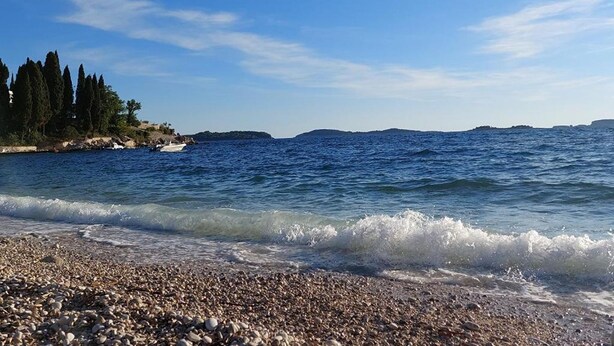 Ein Kiesstrand mit klarem, blauem Wasser und sanften Hügeln im Hintergrund.