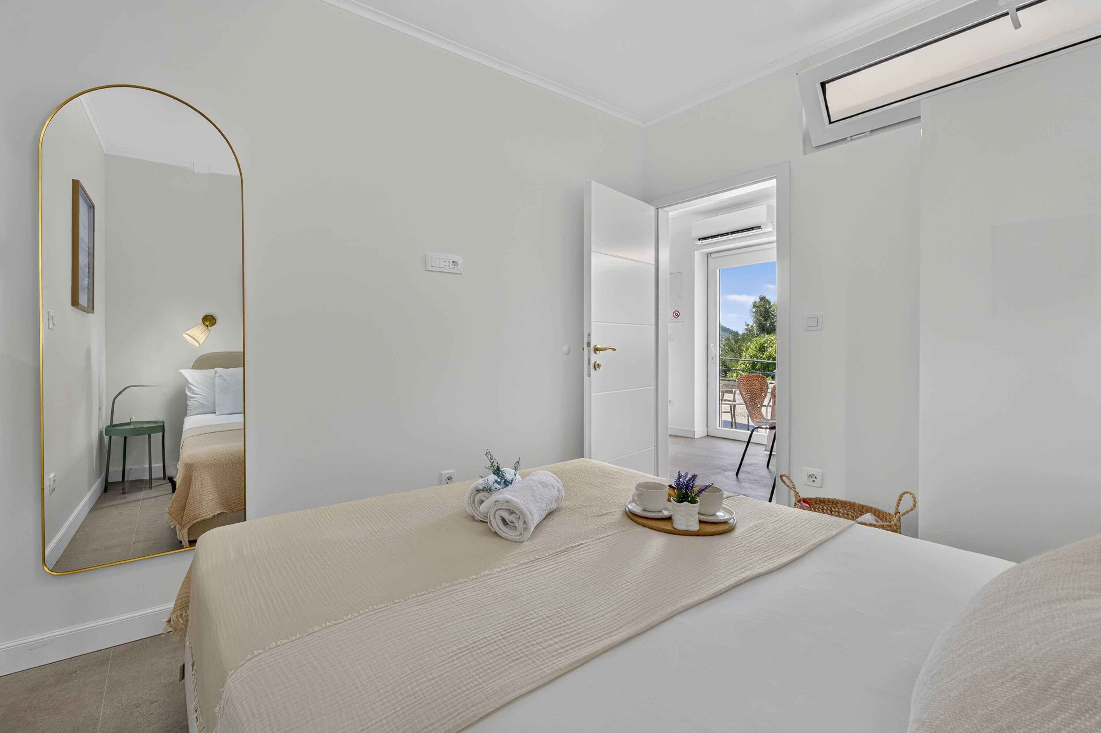 A hotel room with a bed, a mirror, a bedside table with a lamp, a window, and a view of the outdoors.