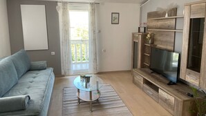A modern living room with a sofa, a glass coffee table, a wooden TV stand, and a balcony with curtains. - Apartmani Meri (Vodice)