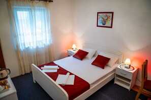 Ein ordentlich bezogenes Bett mit roten Kissen und einer weißen Bettdecke, zwei Nachttische mit Lampen, ein Fenster mit durchscheinenden Vorhängen und ein gerahmtes Bild an der Wand.