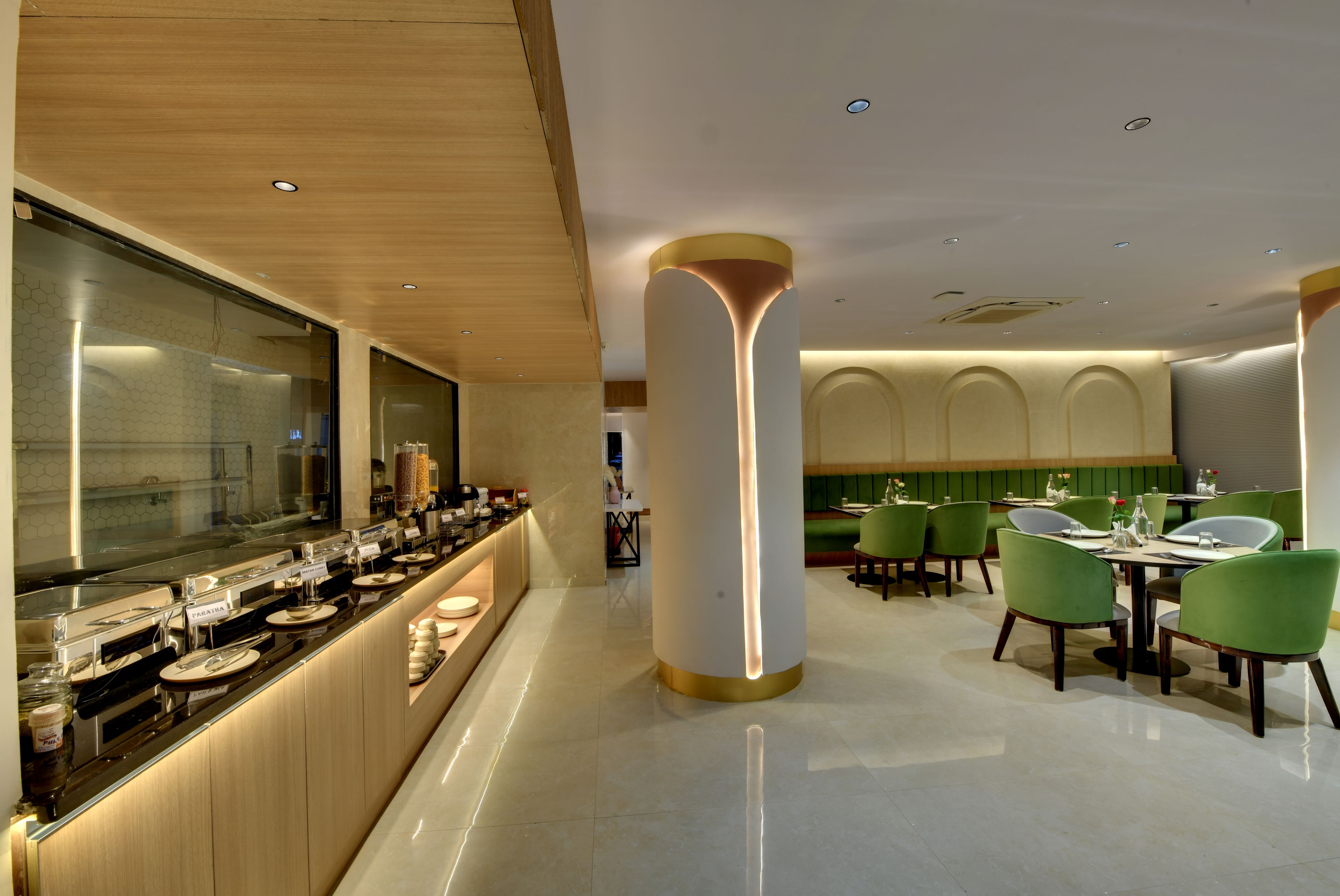Ein modernes Restaurant mit einer langen Theke, grünen Polsterstühlen und einer zentralen Säule mit einer Leuchte.