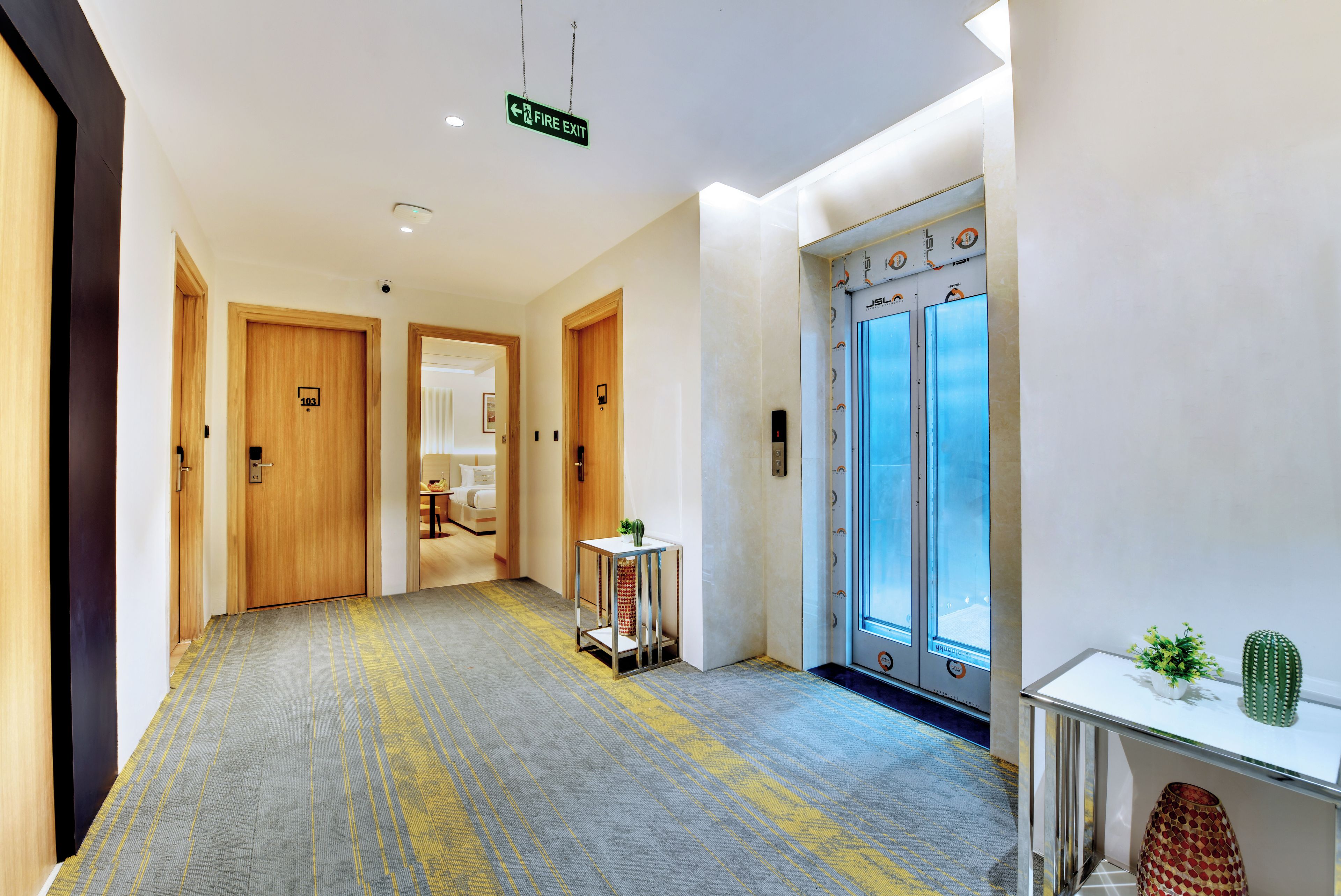 Un pasillo de hotel con puertas de madera, un letrero de salida de incendios, un ascensor y una pequeña mesa con una planta.