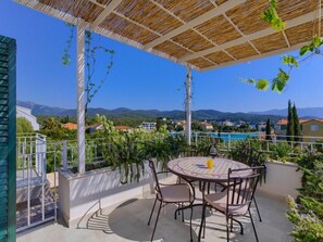 Eine Terrasse im Freien mit einem runden Tisch und Stühlen, umgeben von viel Grün und Blick auf ein Schwimmbecken und Berge.
