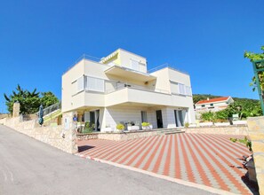 A two-story building with a flat roof, a balcony on the upper floor, and a paved driveway with a red and white pattern. - Apartments Gusti - One Bedroom Apartment With Terrace and Sea View (Janjina)