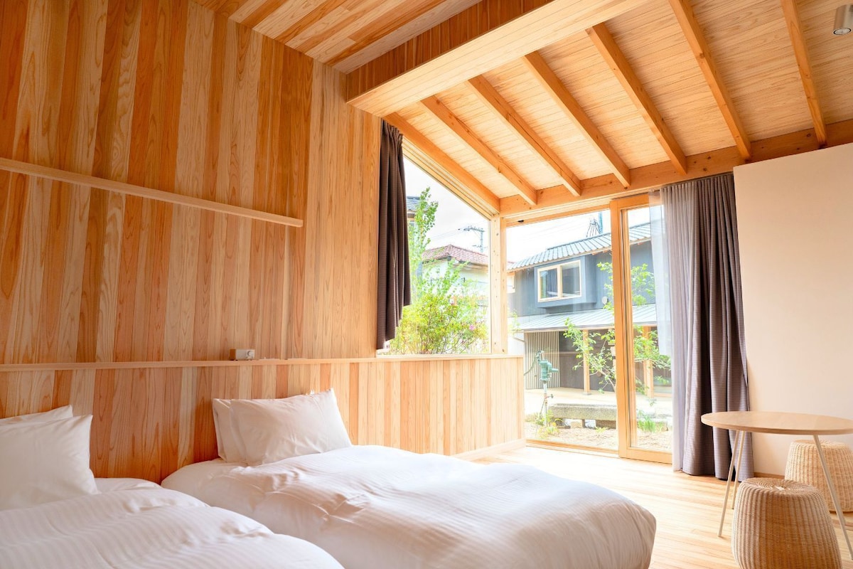 A wooden room with a bed, a small table, and a view of a house through a window.