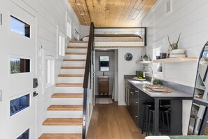 A modern interior with a wooden staircase, a kitchen with bar stools, and a view through a window. - Luxury Tiny House Hot Tub Mountain Views Near Sisters (Sisters)
