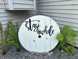 Un panneau circulaire, sur fond blanc, portant l’inscription « Restez un peu », et entouré de petits rochers et de fougères.