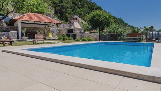 A rectangular swimming pool with a stone wall on one side, a covered seating area with a bench, and a stone building in the background. - Holiday Home Stari Zoganj - Two Bedroom Holiday Home With Terrace and Swimming Pool (Orebic)
