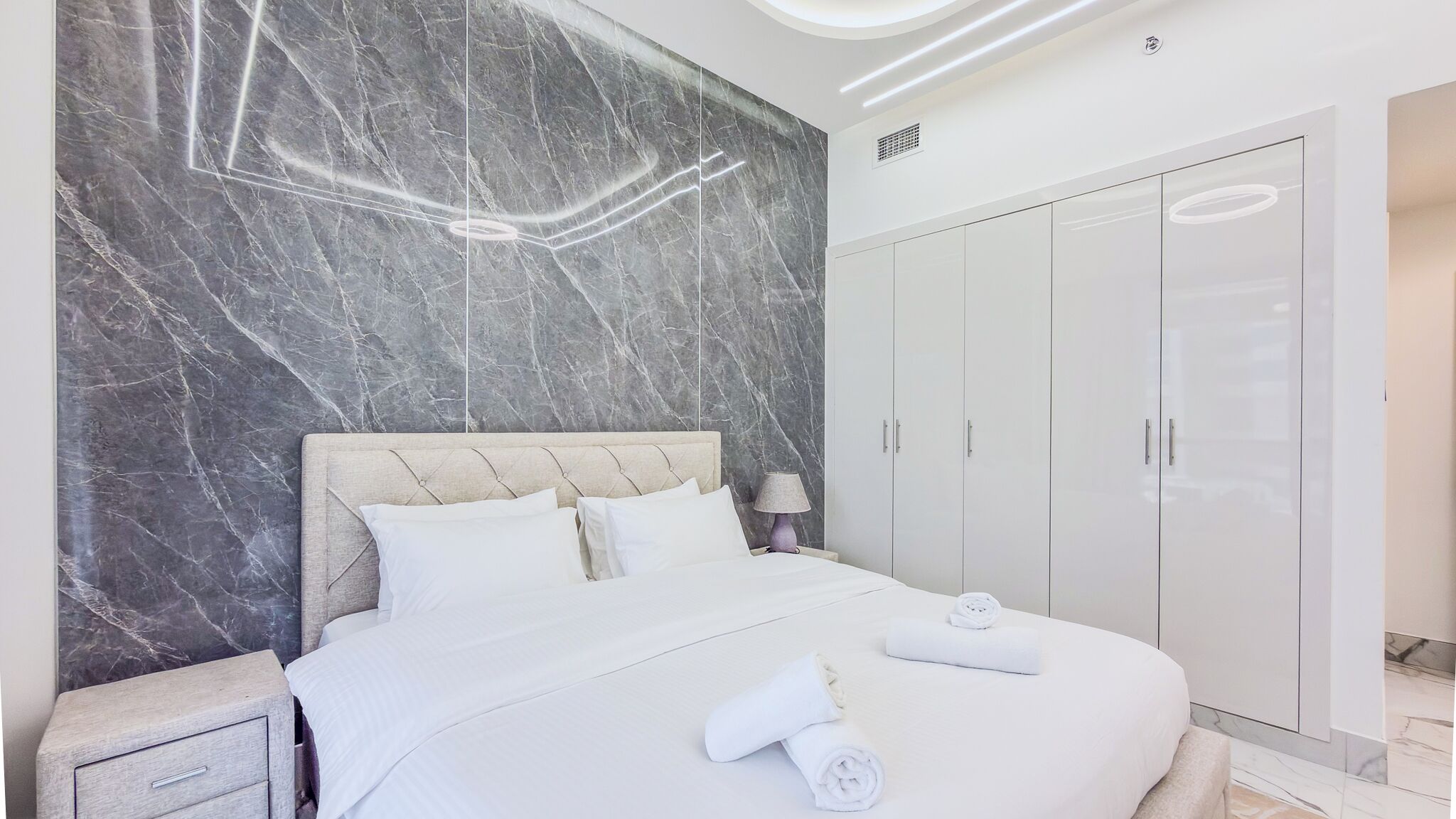 A modern bedroom with a marble wall, a tufted headboard, a large wardrobe, and neatly made towels on the bed.