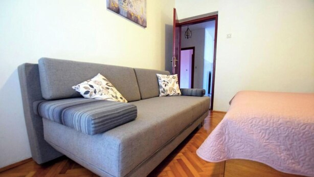 Una habitación con un sofá gris, una cama con manta rosa, suelo de madera y una puerta que da a otra habitación.