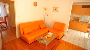 A living room with an orange sofa, a glass coffee table, and a kitchen area with wooden cabinets. - Villa Anni-two Bedroom Apartment With Terrace (Sutivan)