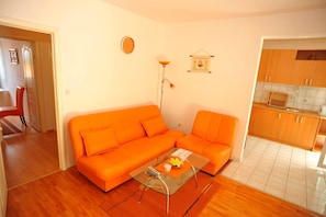 Ein Wohnzimmer mit einem orangefarbenen Sofa, einem Glastisch und einer Küchenzeile mit Holdekor.