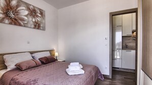 A bedroom with a bed, pillows, a nightstand, a wall art piece, and a view into a kitchen area. - Apartments Victoria - One Bedroom Apartment With Terrace and Sea View (Pakostane)