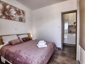 Ein Schlafzimmer mit einem Bett, Kissen, einem Nachttisch, einem Wandbild und Blick in eine Küchenzeile.
