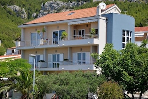 A multi-story building with balconies, a flat roof, and a mountainous backdrop. - Apartments Antonio - One Bedroom Apartment With Balcony - 1 (Tucepi)