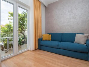 A room with a blue sofa, a yellow cushion, and a view through a glass door to an outdoor area with a table and chairs. - Apartments Antonio - One Bedroom Apartment With Balcony - 1 (Tucepi)