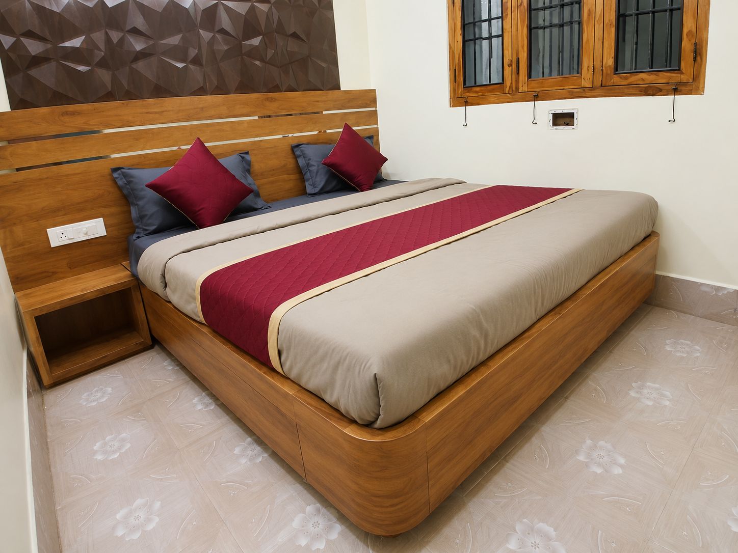 Una cama con cabecera de madera, cubrecama rojo y beige, y una mesita de noche con toma corriente.