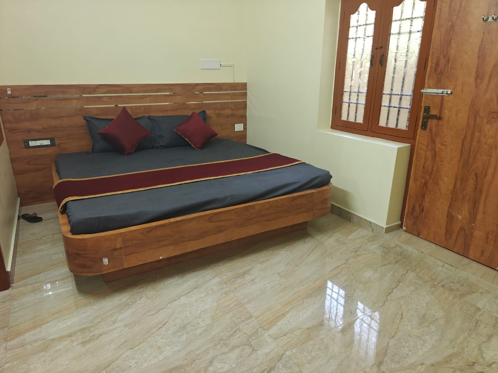 Un dormitorio con cabecera de madera, cama con ropa de cama gris y almohadas rojas, una puerta de madera y una ventana con diseño de cuadrícula.
