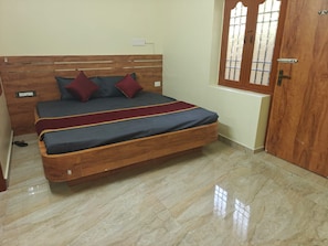 A bedroom with a wooden headboard, a bed with grey bedding and red pillows, a wooden door, and a window with a grid pattern. - Grand Eco Stay (Tiruvannamalai)