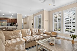 Un salon moderne avec un canapé en cuir beige, une table basse en bois agrémentée d’objets décoratifs et de grandes fenêtres dotées de volets blancs.