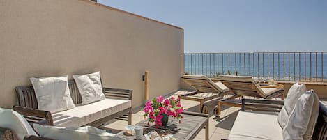 A terrace with a view of the sea, furnished with a sofa, chairs, and a table with flowers.
