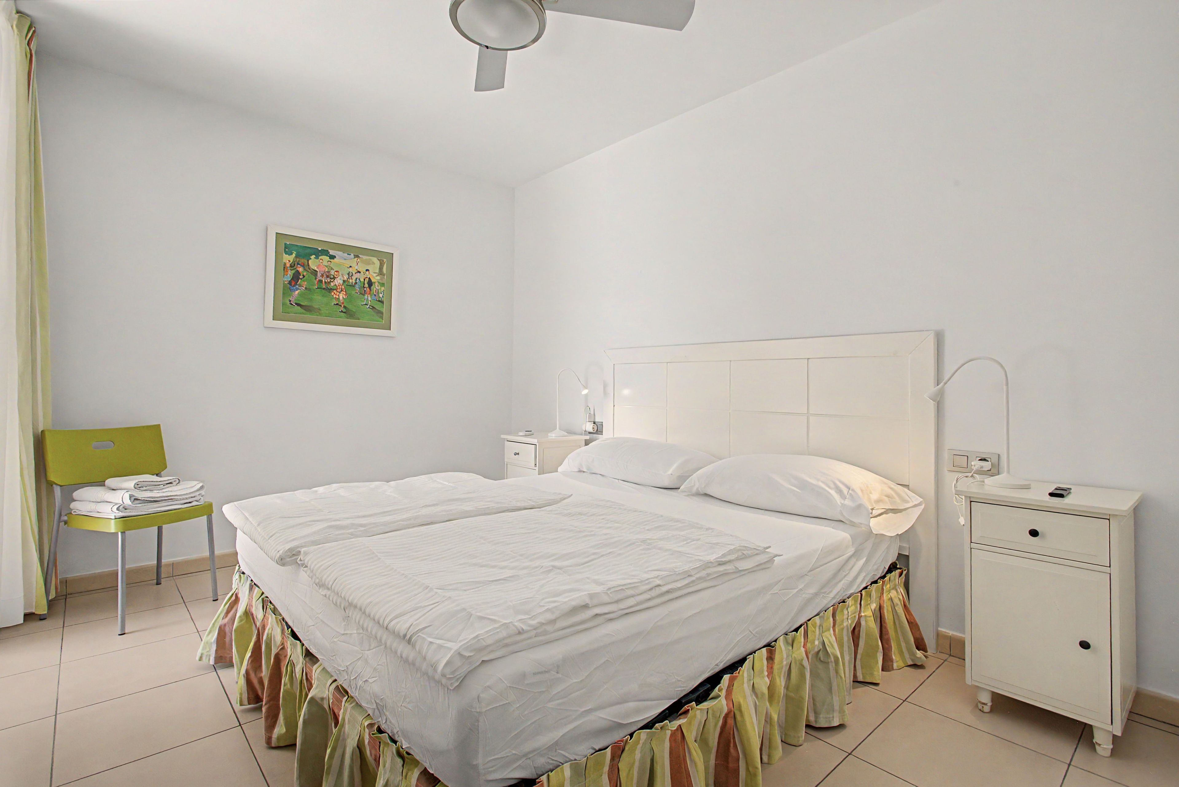 Una cama bien hecha con cabecera blanca, una mesita de noche con lámpara y una silla con toallas dobladas.