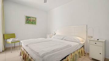 Una cama bien hecha con cabecera blanca, una mesita de noche con lámpara y una silla con toallas dobladas.
