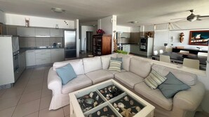 A modern living room with a beige sectional sofa, a glass coffee table with seashells, a kitchen with white cabinets, and a dining area with a table and chairs. - Vacation Home 'Casa Médano 12' with Sea View, Private Terrace and Wi-Fi (El Médano)
