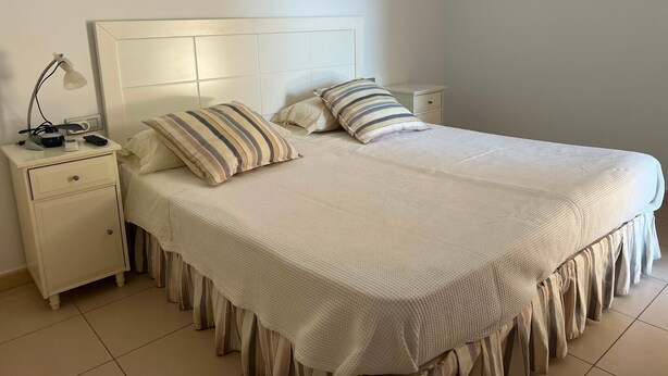 A neatly made bed with a white headboard and a decorative fringe at the foot. There are two bedside tables with lamps and a small drawer. - Vacation Home 'Casa Médano 12' with Sea View, Private Terrace and Wi-Fi (El Médano)