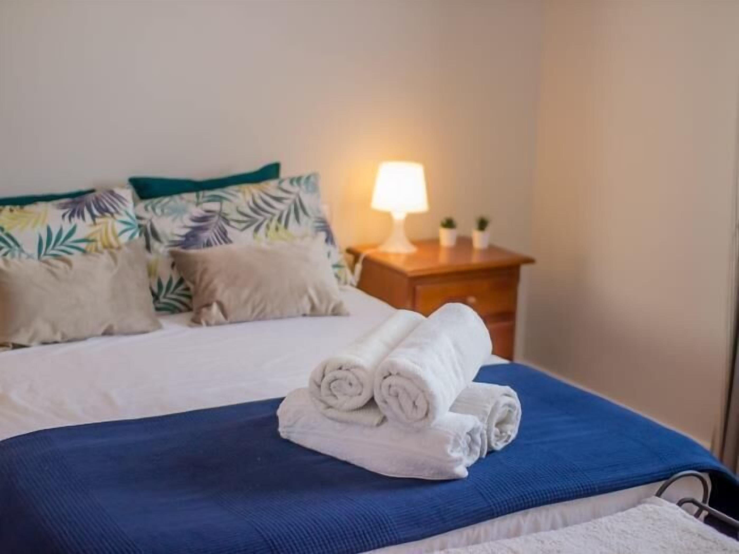 Een net opgemaakt bed met opgerolde handdoeken, een bedlampje en een nachtkastje met een klein plantje.