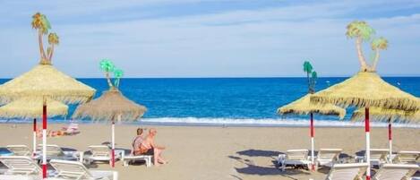 A beach with thatched umbrellas and lounge chairs, a clear sky, and the ocean in the background.