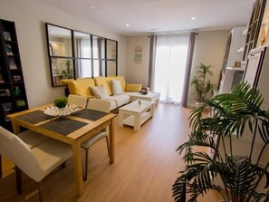 A modern living room with a sofa, coffee table, and dining table. There is a bookshelf and a large mirror on the wall. - Sunstay Flat Torresol (Torremolinos)