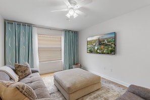 A living room with a grey sofa, a beige ottoman, a ceiling fan, and a cityscape painting on the wall. - Screened Pool + Hot Tub + Game Room | Gated Resort Near Parks | Palm & Play (Kissimmee)