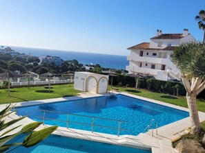 A swimming pool with a clear view of the sea in the background, surrounded by a well-maintained lawn and a white building with a tiled roof. - Sunstay Flat Centauro Mijas (Mijas)
