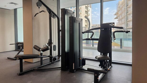 A gym with various exercise equipment, large windows, and a view of buildings outside. - Bloomfields Studio in NAS3 - Arjan Dubai (Dubai)
