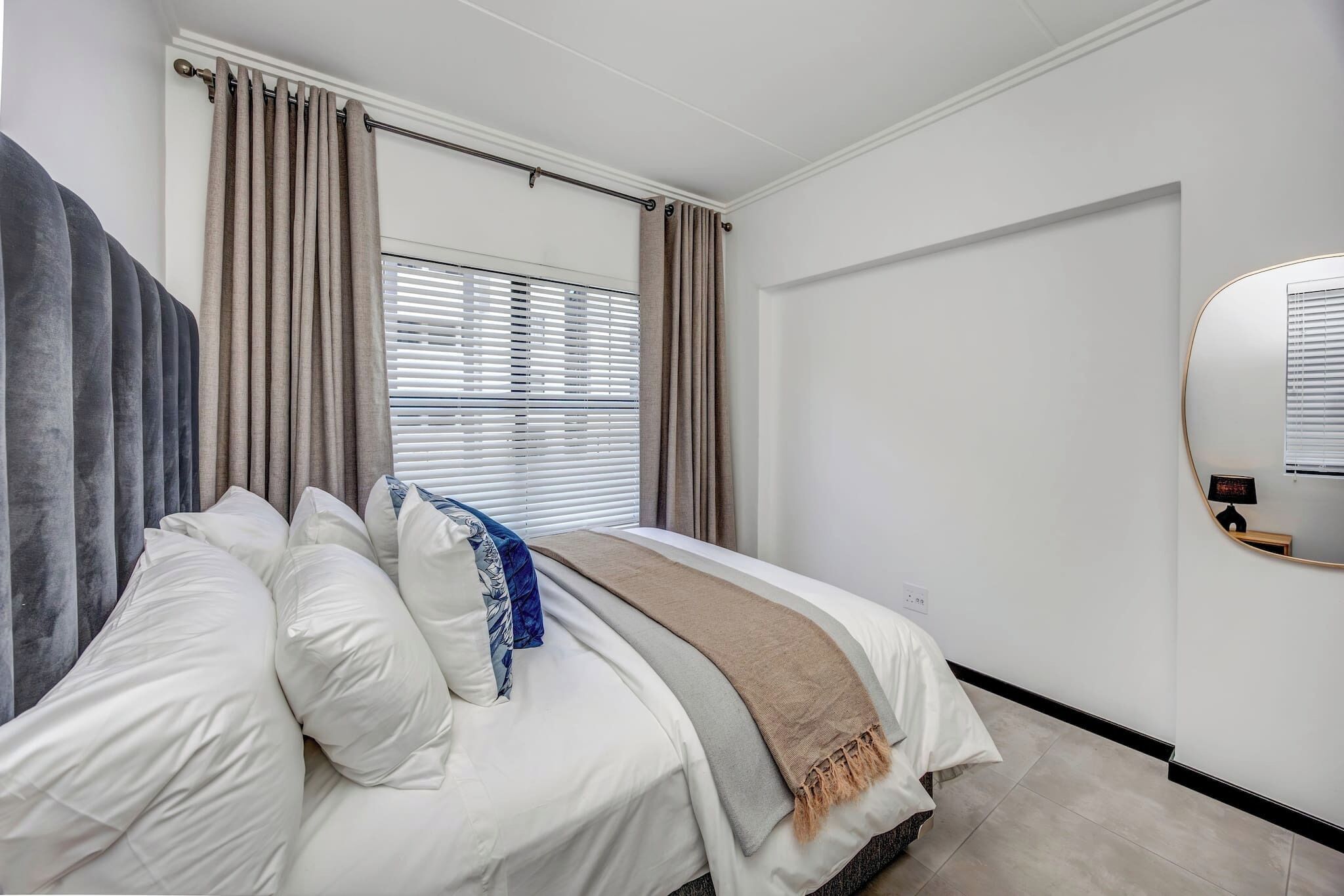 A modern bedroom with a bed, curtains, a window with blinds, a wall-mounted mirror, and a door.