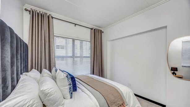 Une chambre à coucher moderne avec un lit, des rideaux, une fenêtre avec des stores, un miroir fixé au mur et une porte.