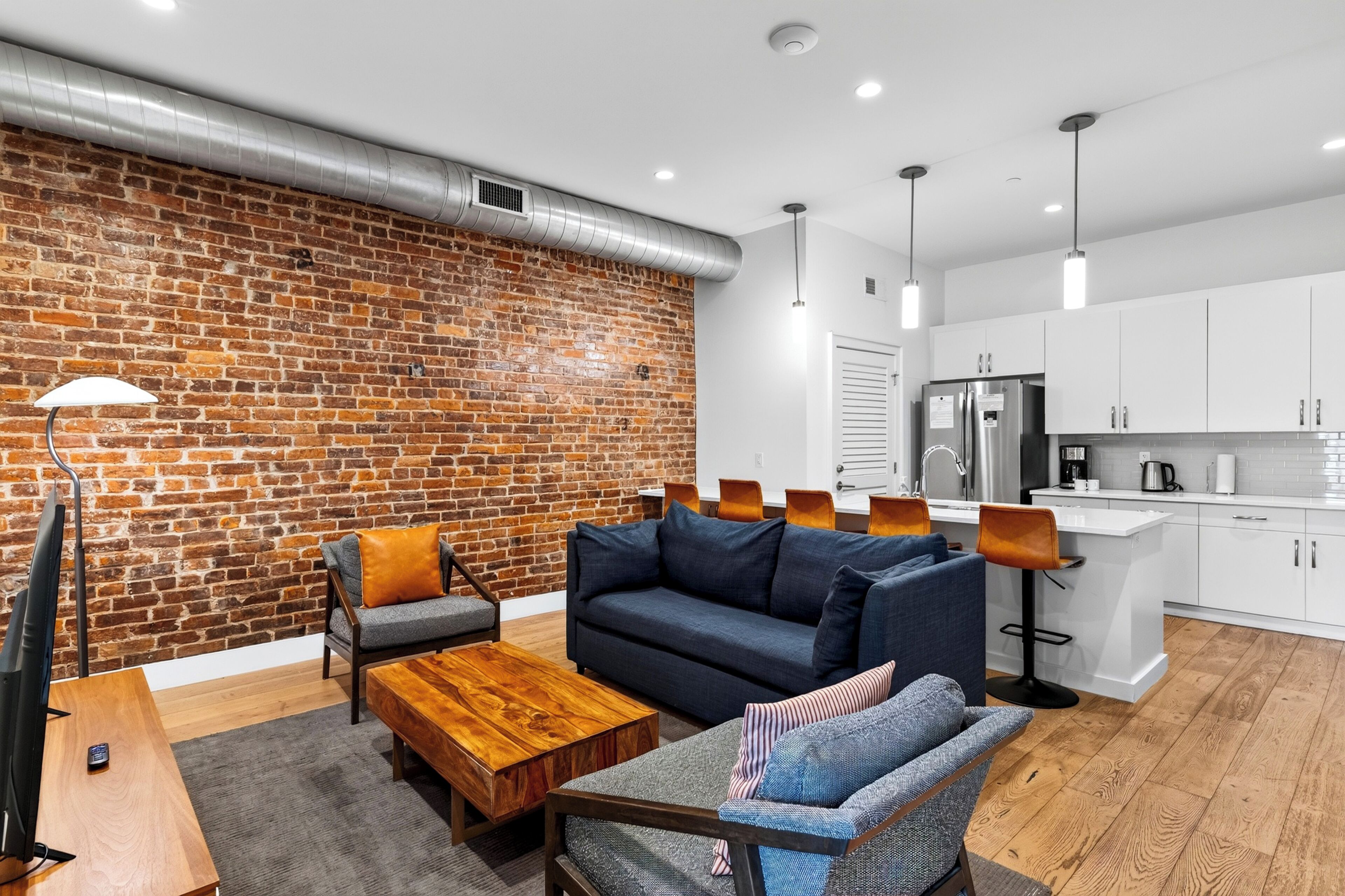 A modern living room with a brick wall, a sofa, a coffee table, and a dining area with a kitchen in the background.