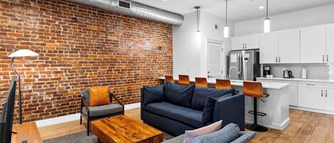 A modern living room with a brick wall, a sofa, a coffee table, and a dining area with a kitchen in the background.