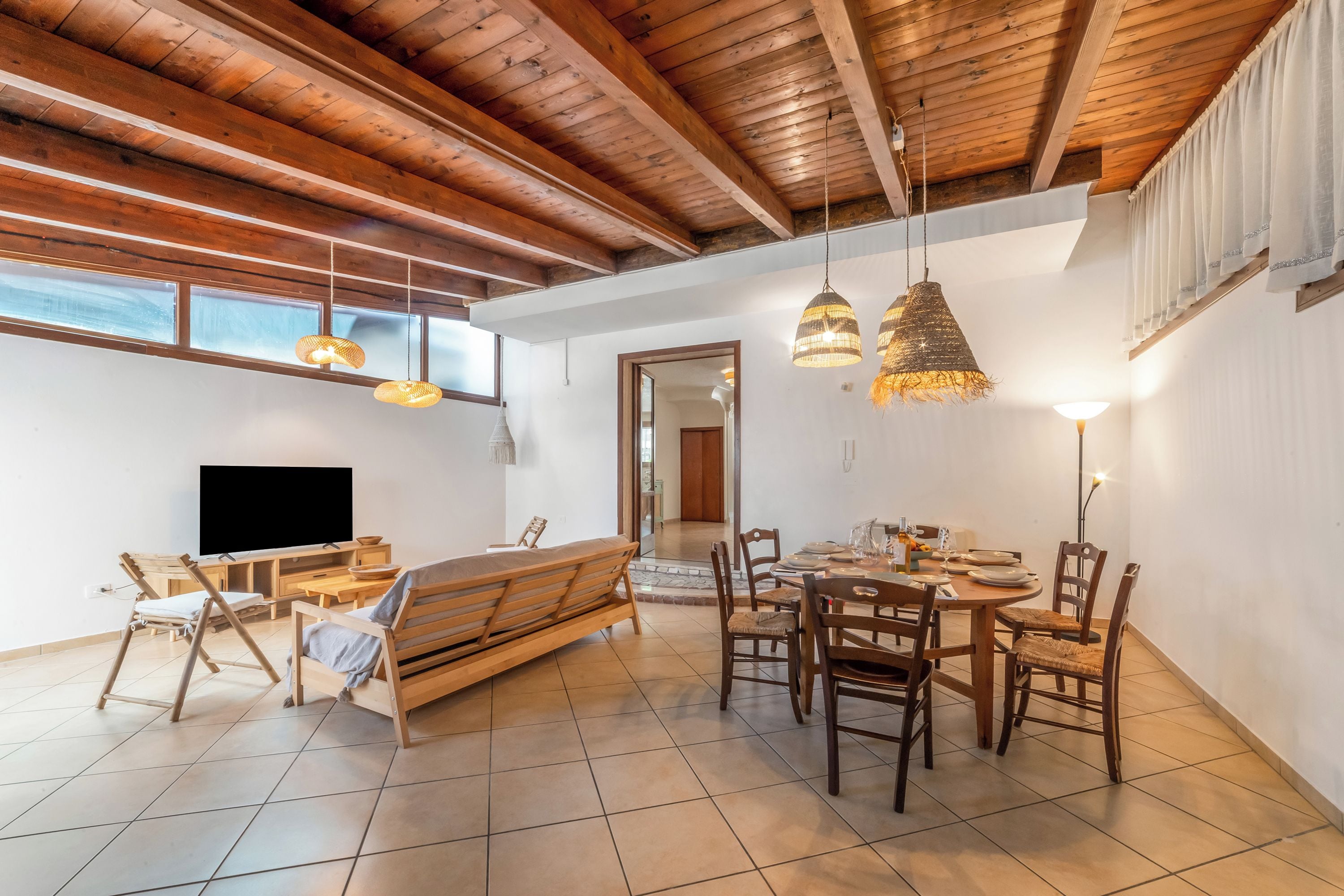 A spacious living area with a wooden ceiling, a TV, a sofa, and a dining table with chairs.