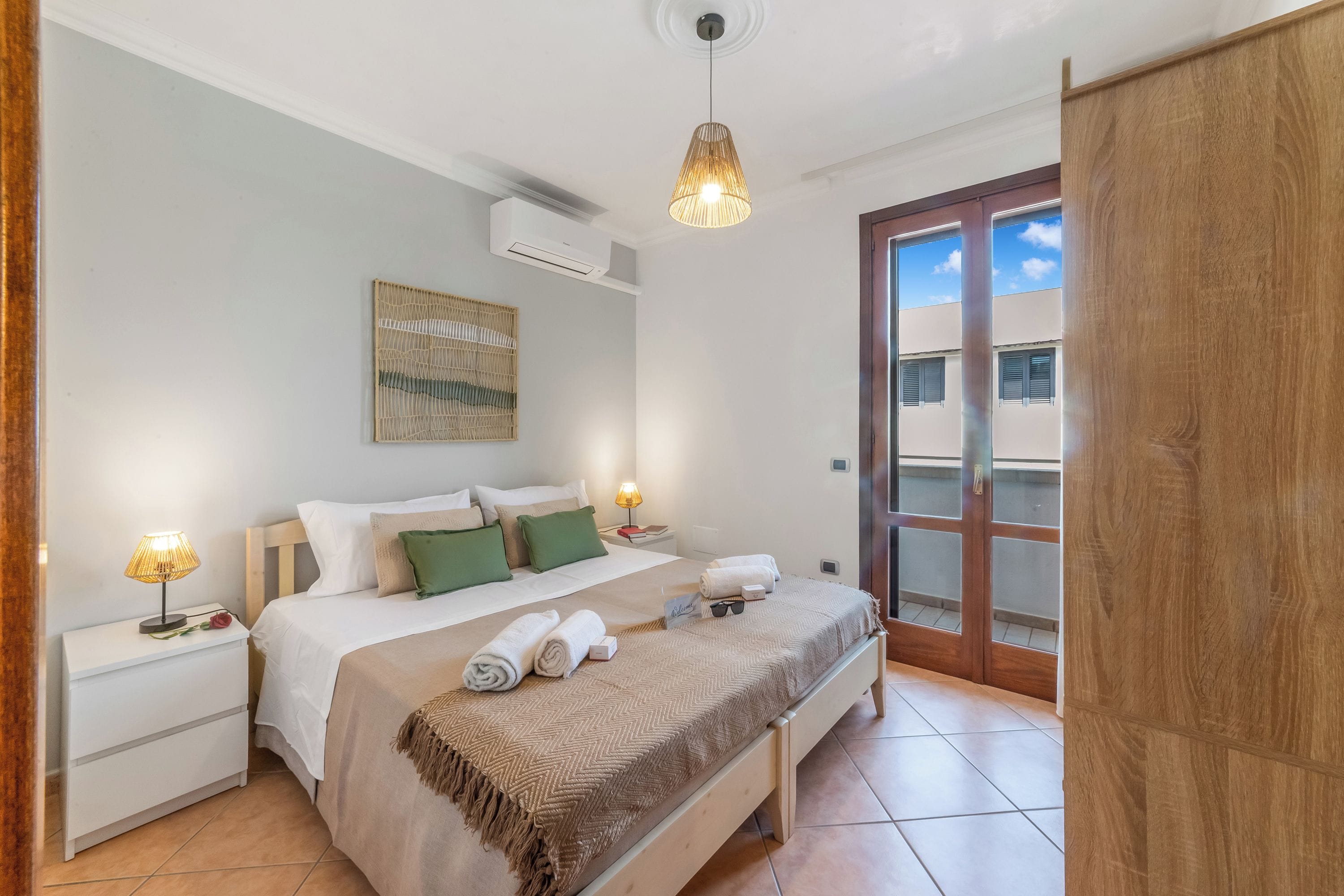 Quarto de hotel com cama, mesas de cabeceira, abajur, uma obra de arte na parede e vista para o exterior por uma porta.
