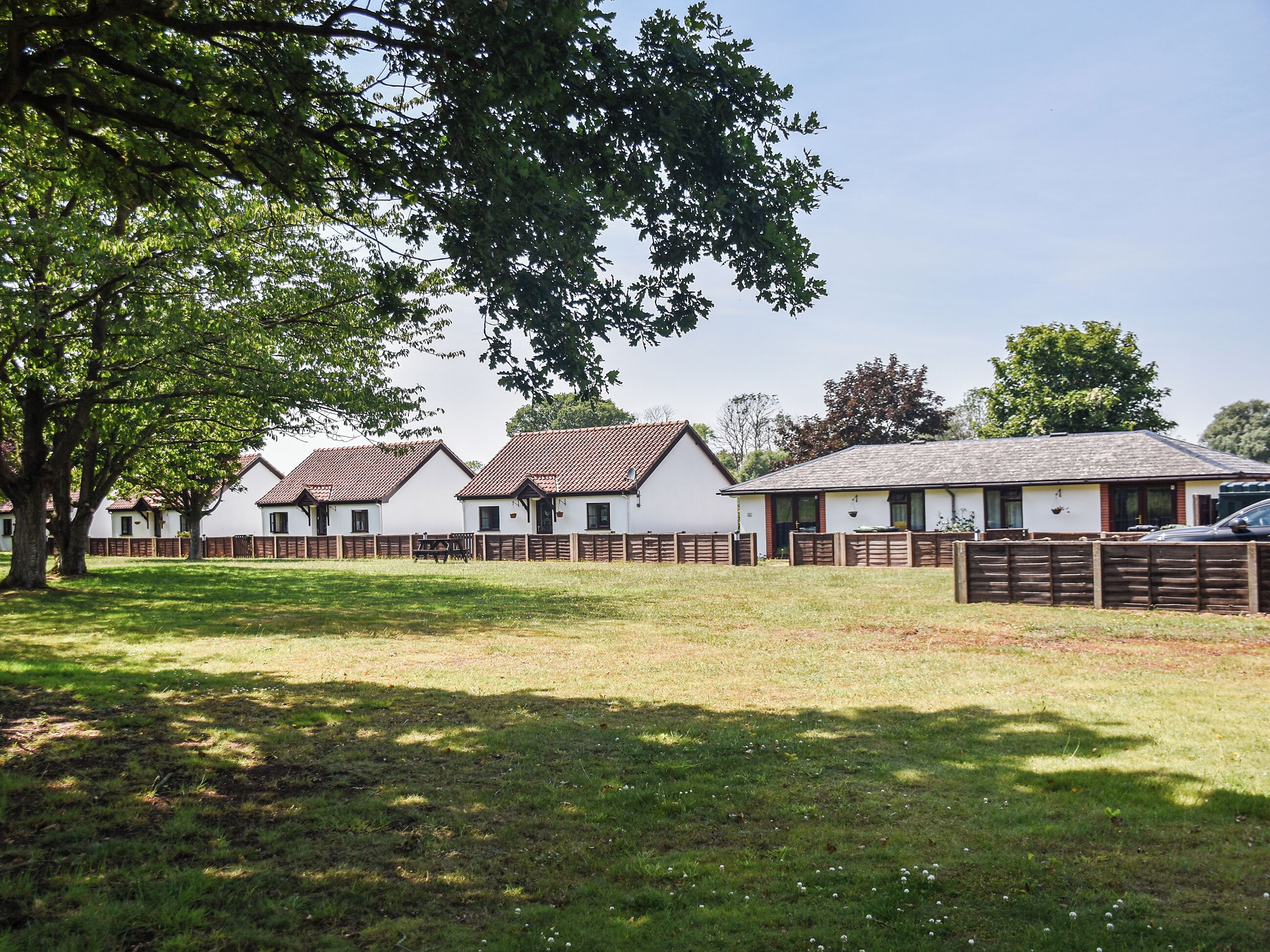 A row of white cottages with brown roofs, surrounded by a wooden fence and a grassy area.